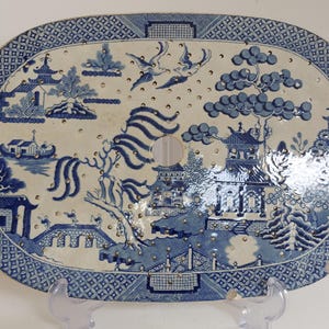 May include: Oval, blue and white ceramic platter with a Willow pattern design. The platter features a central circular opening and is decorated with pagodas, bridges, trees, and birds. The border has a geometric pattern.
