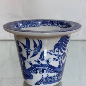 May include: A small, white ceramic planter with a blue Willow pattern design. The planter has a wide, flat rim and a cylindrical body. The blue design depicts birds, a bridge, and other elements. The planter is likely vintage or antique.