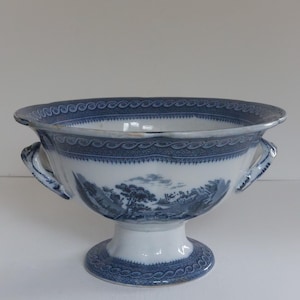 May include: A blue and white porcelain bowl with a pedestal base and two handles. The bowl features a landscape scene and decorative patterns. The rim and base have a repeating wave-like design. The bowl is a decorative piece.