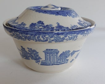 May include: A white ceramic bowl with a lid, featuring a blue and white toile pattern. The bowl has a rounded shape with a lid that has a small knob on top. The blue pattern depicts scenes of buildings and landscapes.