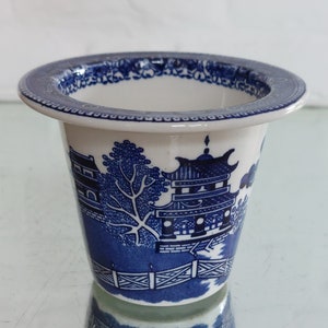 May include: A white and blue ceramic planter with a classic Willow pattern design. The planter features a detailed blue illustration of a pagoda, trees, and a bridge. The rim has a decorative blue border.