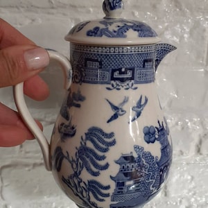 Könnte beinhalten: Eine blau-weiße Keramik-Sahnekännchen mit Deckel. Das Sahnekännchen hat ein Willow-Muster mit Vögeln, Bäumen und Gebäuden. Das Sahnekännchen hat einen Griff und einen Ausguss. Das Sahnekännchen ist etwa 15 cm hoch.