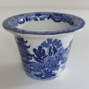 May include: A small, white ceramic planter with a blue Willow pattern design. The planter has a wide, slightly flared rim and a cylindrical body. The blue pattern depicts a landscape with trees, buildings, and a bridge.