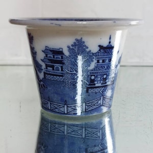 May include: A blue and white ceramic planter with a classic Willow pattern design. The planter features a flared rim and a detailed landscape scene with pagodas and trees. The planter is approximately 4 inches in diameter and 3 inches tall.