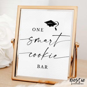 May include: A gold framed sign with the words "ONE smart cookie BAR" in black script. A graduation cap graphic is above the words. The sign is on a light-colored wooden surface.