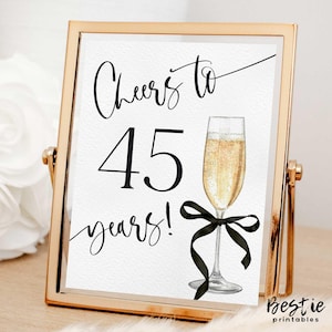 Puede incluir: Un cartel con marco dorado con texto negro que dice "Cheers to 45 years!" con una cinta negra y una copa de champán.
