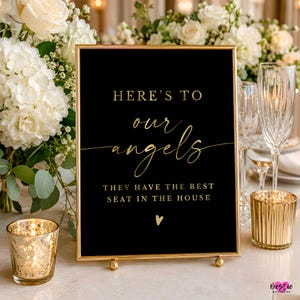 May include: A rectangular black sign in a gold frame featuring gold text that reads: HERE'S TO our angels THEY HAVE THE BEST SEAT IN THE HOUSE, with a small gold heart icon at the bottom. The sign is displayed upright on a flat surface.