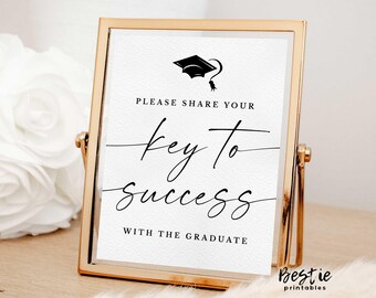 May include: A gold-framed sign with a white background and black text. The sign reads "Please share your key to success with the graduate". A black graduation cap is at the top of the sign.