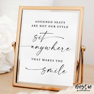May include: A gold-framed sign with the text "Assigned seats are not our style. Sit anywhere that makes you smile."  The sign is sitting on a white surface.