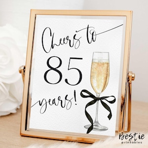May include: Gold-framed sign with black text that reads "Cheers to 85 years!" with a black ribbon and a glass of champagne.