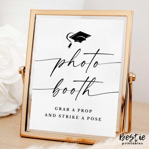 May include: A gold-framed sign with the words "photo booth" in elegant script, topped with a graduation cap illustration. The text "Grab a prop and strike a pose" is below. The sign is on a wooden surface.