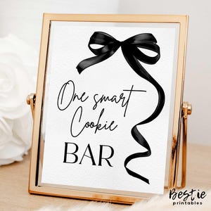 以下が含まれることがあります： 黒いリボンボウと「One smart cookie BAR」という黒い筆記体で書かれたテキストが書かれた金色の額縁の看板です。
