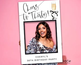 Op de afbeelding: Een witte fotobooth-prop met een zwart frame en de tekst "Cheers to Thirty!" Een glas champagne met een zwarte strik bevindt zich rechtsboven. De tekst "Cheryl's 30th Birthday Party" staat onderaan de prop.