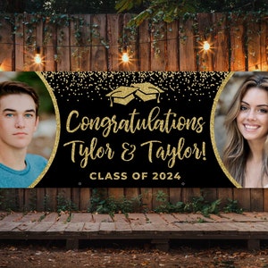 May include: A black banner with gold glitter accents and the text "Congratulations Tylor & Taylor! CLASS OF 2024." Features two photos, one of a young man and one of a young woman. The banner is displayed outdoors against a wooden backdrop.