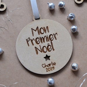 K&ouml;nnte beinhalten: Ein runder Holz-Anh&auml;nger mit dem Text "Mon Premier No&euml;l" und "Charlie 2019" eingraviert. Der Anh&auml;nger ist mit einem kleinen Stern verziert.
