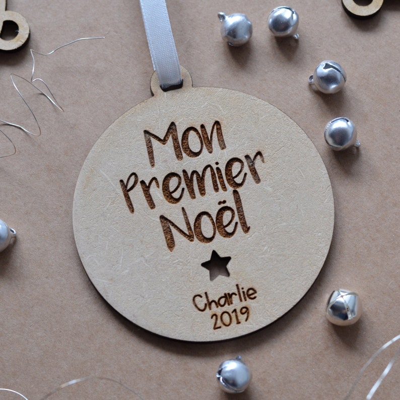 K&ouml;nnte beinhalten: Ein runder Holz-Anh&auml;nger mit dem Text "Mon Premier No&euml;l" und "Charlie 2019" eingraviert. Der Anh&auml;nger ist mit einem kleinen Stern verziert.