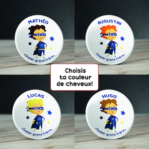 May include: Four white round buttons with blue and yellow superhero designs. Each button features a different name: Mathéo, Augustin, Lucas, and Hugo. The text "Super grand frère" is printed on each button. The buttons are on a wooden surface.
