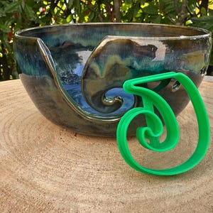 Puede incluir: Un bol de lana de cerámica azul oscuro y verde con un guía de lana de plástico verde. El bol tiene un diseño de espiral único y es perfecto para guardar lana mientras se teje o se hace ganchillo.