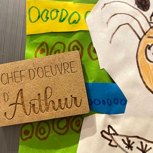 May include: A wooden plaque with the text "Chef d'oeuvre d'Arthur" engraved on it. The plaque is brown and has a rough texture.