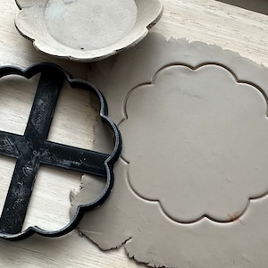 May include: A black plastic cookie cutter in a scalloped flower shape, next to a cut-out of the same shape in gray clay. A small, unglazed ceramic dish in the same shape is also present. The items are on a light wood surface.