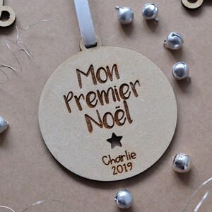 K&ouml;nnte beinhalten: Ein h&ouml;lzerner Weihnachtsbaumschmuck mit dem Text "Mon Premier No&euml;l" und dem Namen "Charlie" sowie dem Jahr "2019" eingraviert. Der Schmuck ist mit einem Stern verziert und hat ein wei&szlig;es Band.