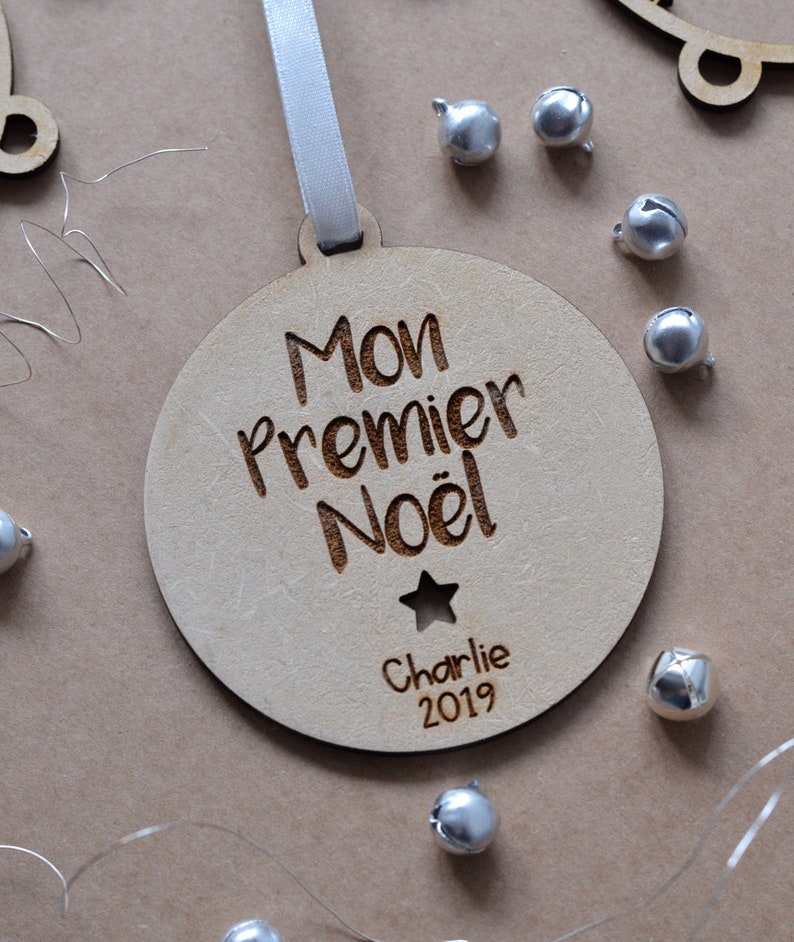 K&ouml;nnte beinhalten: Ein h&ouml;lzerner Weihnachtsbaumschmuck mit dem Text "Mon Premier No&euml;l" und dem Namen "Charlie" sowie dem Jahr "2019" eingraviert. Der Schmuck ist mit einem Stern verziert und hat ein wei&szlig;es Band.