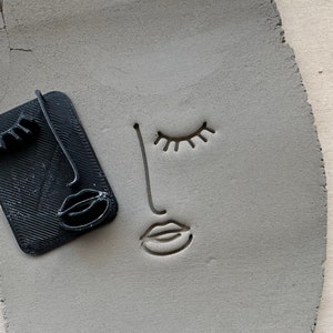 Peut inclure: Un moule en plastique noir d'un visage stylisé avec les yeux fermés, un nez et une bouche. Le moule est utilisé pour créer une empreinte dans de l'argile grise.