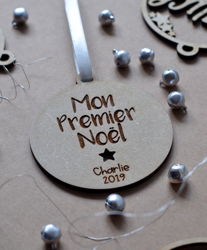 K&ouml;nnte beinhalten: Ein h&ouml;lzerner Weihnachtsbaumschmuck mit dem Text "Mon Premier Noel" und dem Namen "Charlie" sowie dem Jahr "2019" eingraviert. Der Schmuck ist mit einem kleinen Stern verziert und h&auml;ngt an einem wei&szlig;en Band.