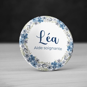 Peut inclure: Un bouton rond blanc orné d'un motif floral et du prénom "Léa" en écriture bleu foncé. Le bouton est bordé de fleurs bleues et de feuilles vertes, avec le texte "Aide soignante" en dessous.
