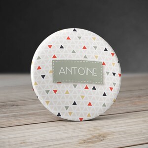 Puede incluir: Un botón redondo con fondo blanco y un patrón geométrico de triángulos negros, rojos y amarillos. El botón tiene una etiqueta verde con el nombre "ANTOINE" en texto blanco.