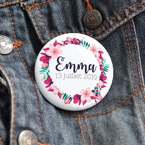 Op de afbeelding: Een witte knop met een ontwerp van een bloemenkrans. De krans is gemaakt van roze en groene bloemen. De tekst "Emma 13 juillet 2019" staat in het midden van de knop.