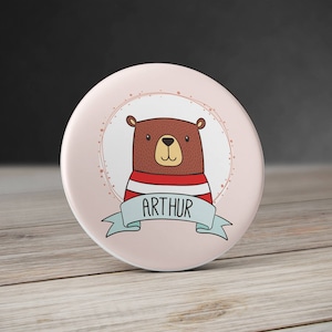 Pode incluir: Um botão redondo rosa com um urso marrom vestindo uma camisa listrada vermelha e branca. O urso está dentro de um círculo com pontos brancos. O texto "ARTHUR" está em uma fita azul abaixo do urso.