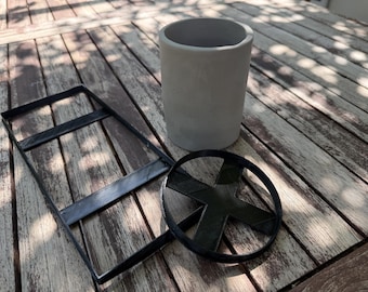 May include: A light gray cylindrical container with a smooth matte finish, accompanied by a black rectangular frame with two crossbars and a separate black circular ring insert featuring a central cross-shaped support structure.