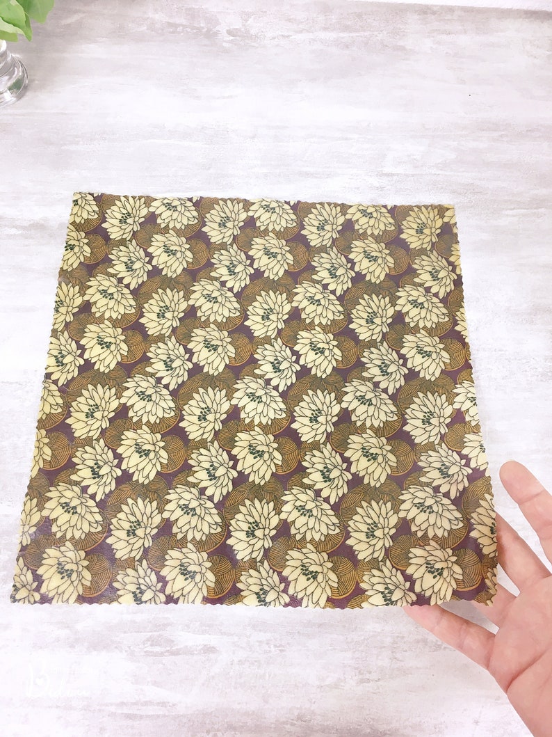 May include: A square piece of beeswax food wrap with a floral pattern. The pattern is yellow and brown on a burgundy background. The wrap is made of natural materials and is reusable and compostable.