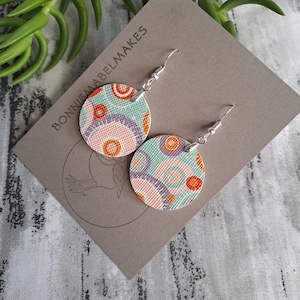 May include: A pair of round, colourful earrings with a geometric pattern. The earrings are made of fabric and have silver hooks. The fabric is a light blue with orange, pink, and purple circles.