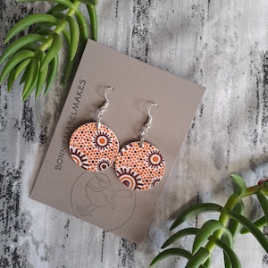 May include: A pair of round earrings with a white background and orange and brown abstract pattern. The earrings are on a brown card with the text "BONNIEMABELMAKES" printed on it.