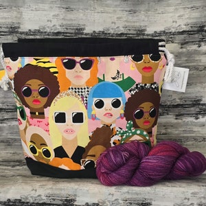 May include: A black drawstring bag featuring a colourful print of women wearing sunglasses. The bag is filled with purple yarn.