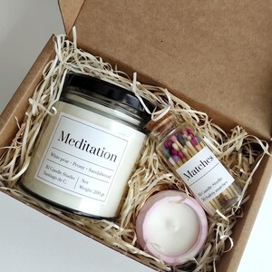 Könnte beinhalten: Eine Geschenkbox gefüllt mit braunem Packpapier und zwei Kerzen. Die größere Kerze befindet sich in einem durchsichtigen Glasgefäß mit schwarzem Deckel und einem weißen Etikett mit der Aufschrift "Meditation White pear - Peony - Sandalwood BJ Candle Studio Santiago de C. Net Weight: 200 g.". Die kleinere Kerze befindet sich in einem rosa Behälter. Ein kleines Glasgefäß mit einem Korkverschluss enthält bunte Streichhölzer. Das Etikett auf dem Glasgefäß lautet "Matches Bl Candle Studio 50 Safety matches".