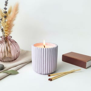 Peut inclure: Une bougie lavande allumée dans un contenant en béton cannelé. La bougie est posée sur une surface blanche avec une boîte d'allumettes et des allumettes. Un petit vase rose avec des fleurs séchées est en arrière-plan.