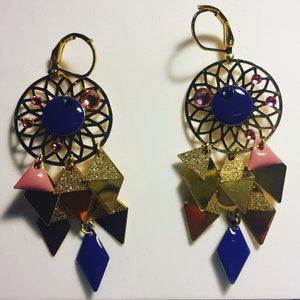 May include: A pair of gold earrings with a blue and pink geometric design. The earrings feature a circular design with a blue center and pink accents. The earrings are adorned with gold and pink geometric shapes.