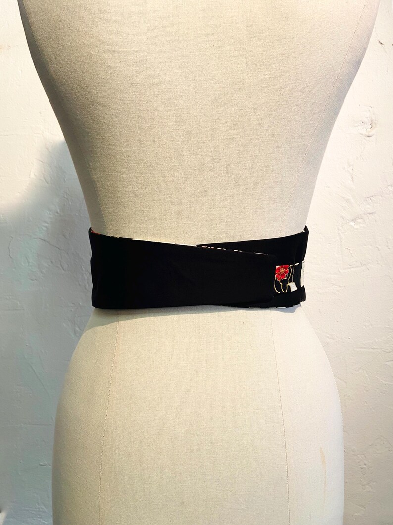 May include: A black obi belt with a red and white floral design. The belt is wrapped around a mannequin. The belt is made of a thick, sturdy material and is designed to cinch the waist.