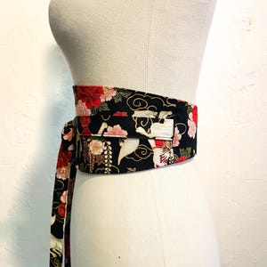 May include: A black obi belt with a floral and crane pattern. The belt features red, pink, and white flowers, with a design of cranes and clouds. The belt is tied around a mannequin, showcasing its wrap-around style.