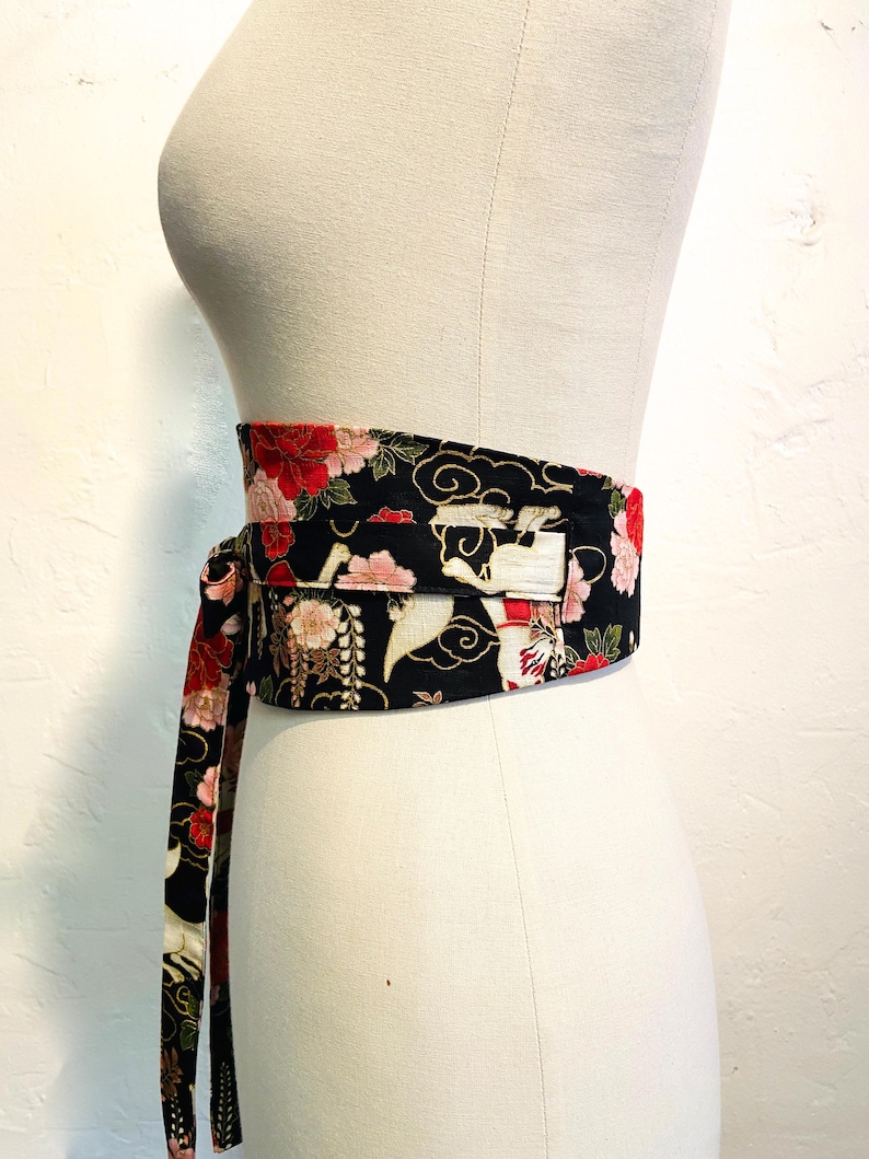 May include: A black obi belt with a floral and crane pattern. The belt features red, pink, and white flowers, with a design of cranes and clouds. The belt is tied around a mannequin, showcasing its wrap-around style.