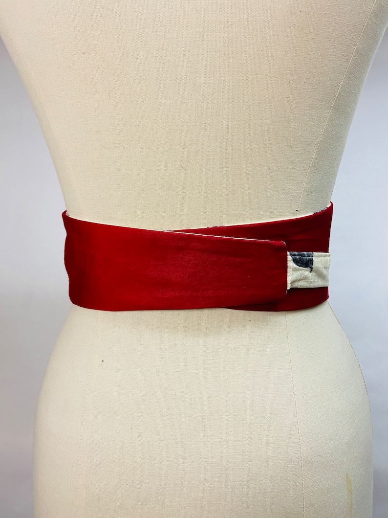 Op de afbeelding: Een brede wikkelriem met een buitenkant van effen dieprode stof die overlapt en een contrasterende gebroken witte voering met een donker, abstract patroon onthult. De riem is horizontaal bevestigd rond een torso van een etalagepop.