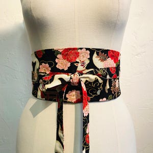 May include: A black obi belt with a floral and crane pattern. The belt features red and pink flowers, white cranes, and gold accents. The belt is tied in a bow in the front, and is displayed on a mannequin.