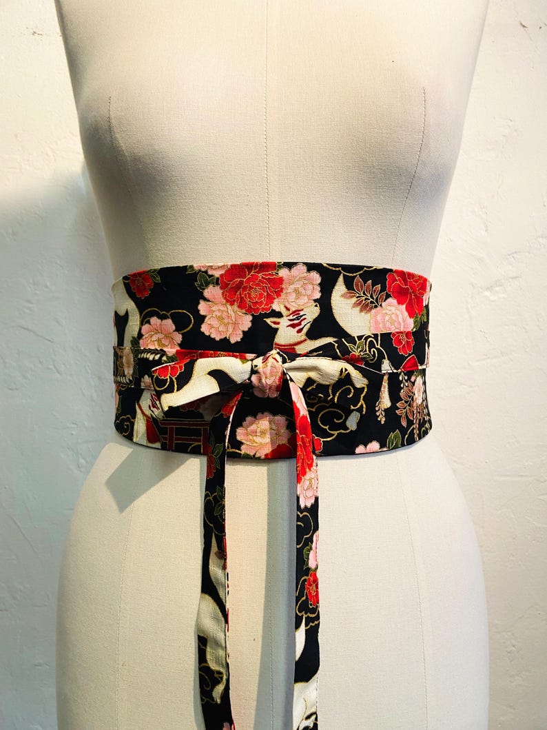 May include: A black obi belt with a floral and crane pattern. The belt features red and pink flowers, white cranes, and gold accents. The belt is tied in a bow in the front, and is displayed on a mannequin.