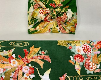 Könnte beinhalten: Ein grünes Furoshiki-Tuch, ein traditionelles japanisches Verpackungstuch, verknotet. Der Stoff zeigt ein Blumen- und Kranichdesign in Rot, Gold und Weiß. Der untere Teil des Bildes zeigt einen passenden Stoff.