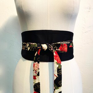 May include: A black obi belt with a floral pattern in red, pink, and white. The belt is tied in a bow, with the patterned fabric hanging down. The belt is displayed on a mannequin.
