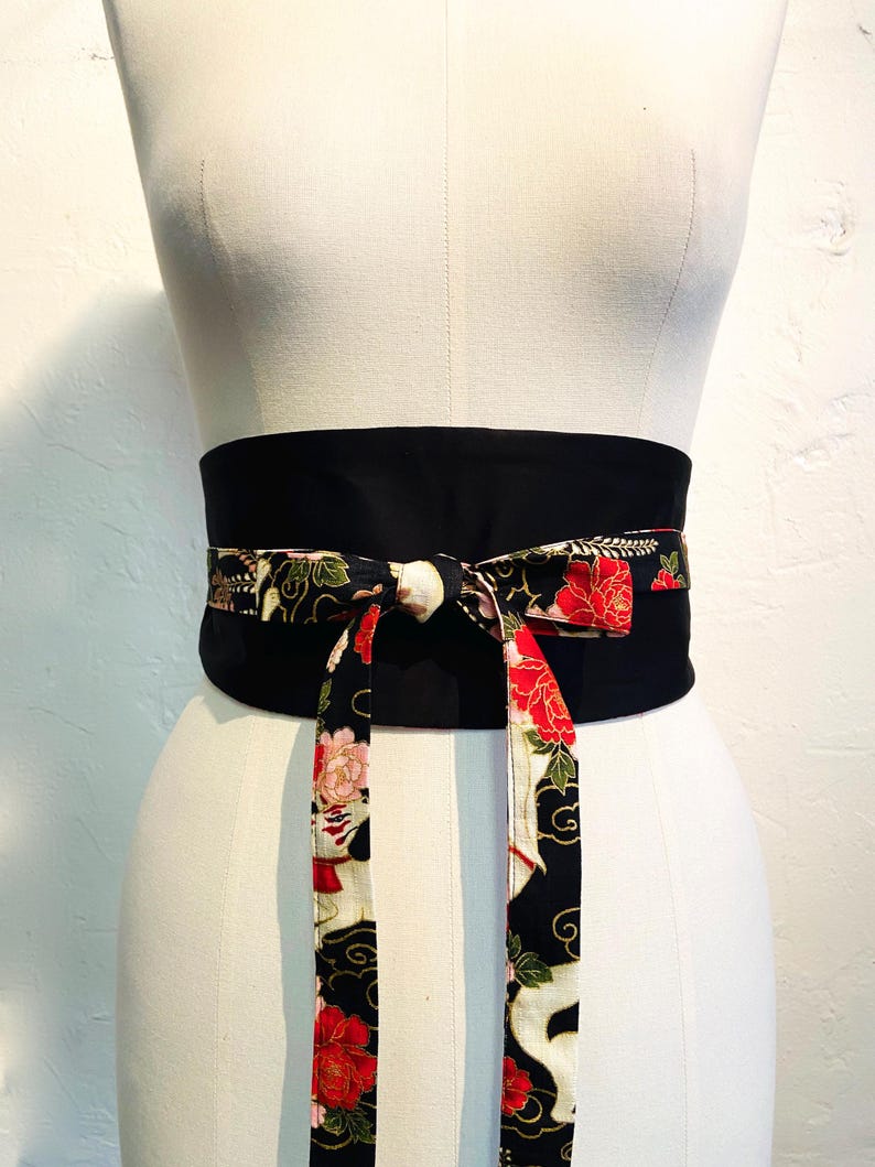 May include: A black obi belt with a floral pattern in red, pink, and white. The belt is tied in a bow, with the patterned fabric hanging down. The belt is displayed on a mannequin.