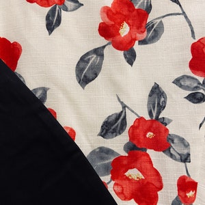 Op de afbeelding: Een textielstof met een cr&egrave;mekleurige achtergrond en een herhalend patroon van felrode bloemen en donkergrijze bladeren. De stof heeft een zichtbare geweven textuur en wordt gedeeltelijk overlapt door een effen, glad, matzwart stoffen gedeelte.